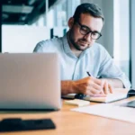 Hombre trabajando con una laptop mientras anota en una libreta, en una oficina con ventanales