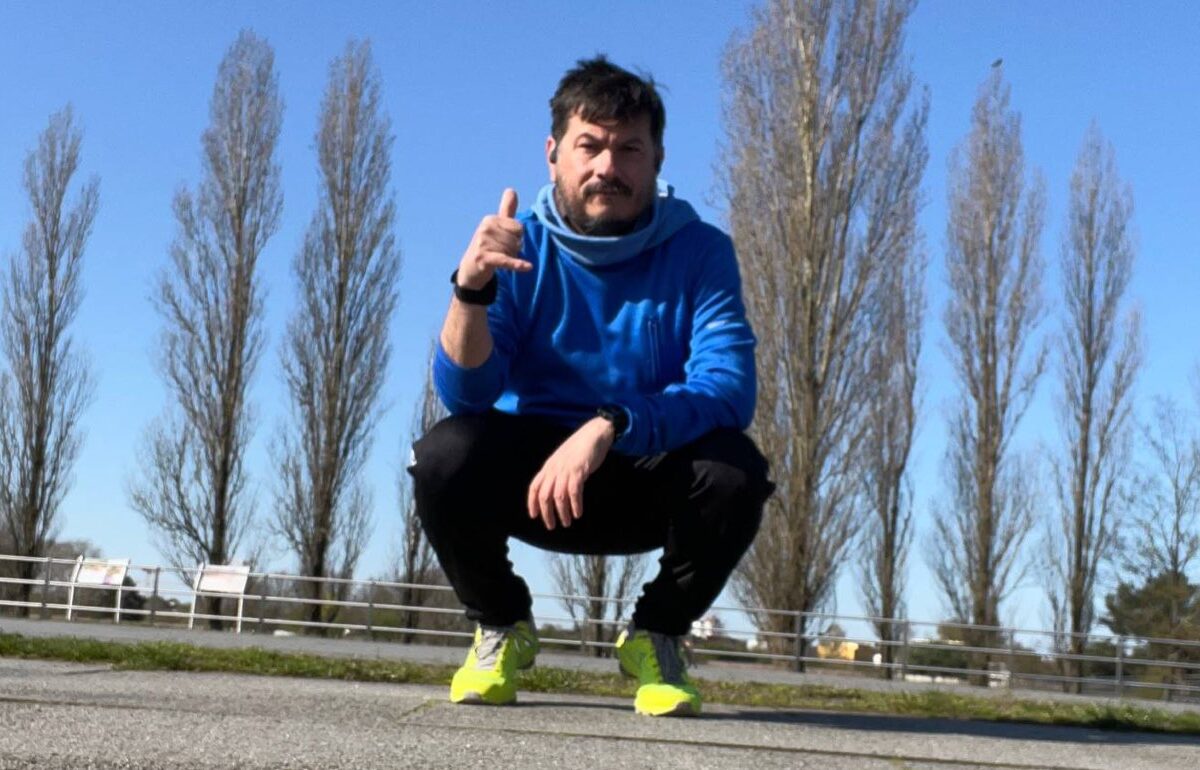 Victor Alvarado en cuclillas sobre una pista de asfalto, con ropa para correr y tenis amarillos, frente a una fila de álamos.