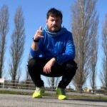 Victor Alvarado en cuclillas sobre una pista de asfalto, con ropa para correr y tenis amarillos, frente a una fila de álamos.
